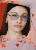 A woman with straight dark hair poses with one arm raised against a red background, wearing the Evangeline blue-tinted aviator sunglasses with a stainless steel frame, silver hoop earrings, and a pink patterned top.