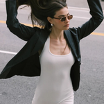 Wearing the Evangeline aviator sunglasses with a stainless steel frame, a woman in a black blazer and white romper flips her long hair with both hands while walking on a city street.