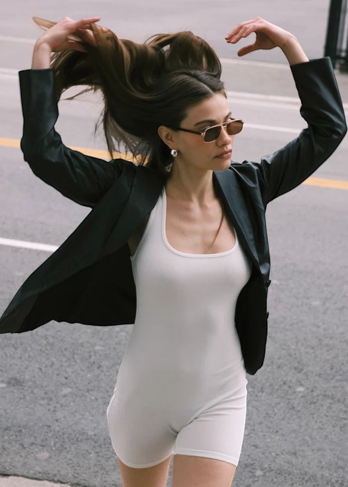 Wearing the Evangeline aviator sunglasses with a stainless steel frame, a woman in a black blazer and white romper flips her long hair with both hands while walking on a city street.