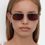 A woman with straight, light brown hair wears the Evangeline dark rectangular sunglasses, which offer UV protection, and small hoop earrings. She looks directly at the camera with a neutral expression, dressed in a white top against a plain background.