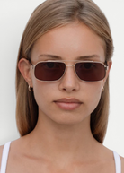 A woman with straight, light brown hair wears the Evangeline dark rectangular sunglasses, which offer UV protection, and small hoop earrings. She looks directly at the camera with a neutral expression, dressed in a white top against a plain background.