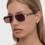 A woman with straight, light brown hair wears the Evangeline rectangular brown sunglasses, which feature a stainless steel frame and is styled with a small gold hoop earring. She has a neutral expression and is dressed in a white top against a plain background.