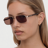 A woman with straight, light brown hair wears the Evangeline rectangular brown sunglasses, which feature a stainless steel frame and is styled with a small gold hoop earring. She has a neutral expression and is dressed in a white top against a plain background.