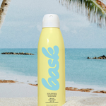 KABE SHOP INC yellow continuous spray sunscreen bottle on sandy beach with palm tree and ocean background