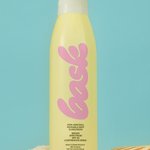 KABE SHOP INC SPF 50 sunscreen spray in a yellow bottle with pink branding, standing on sand against a blue background.
