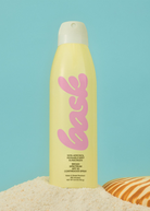 KABE SHOP INC SPF 50 sunscreen spray in a yellow bottle with pink branding, standing on sand against a blue background.