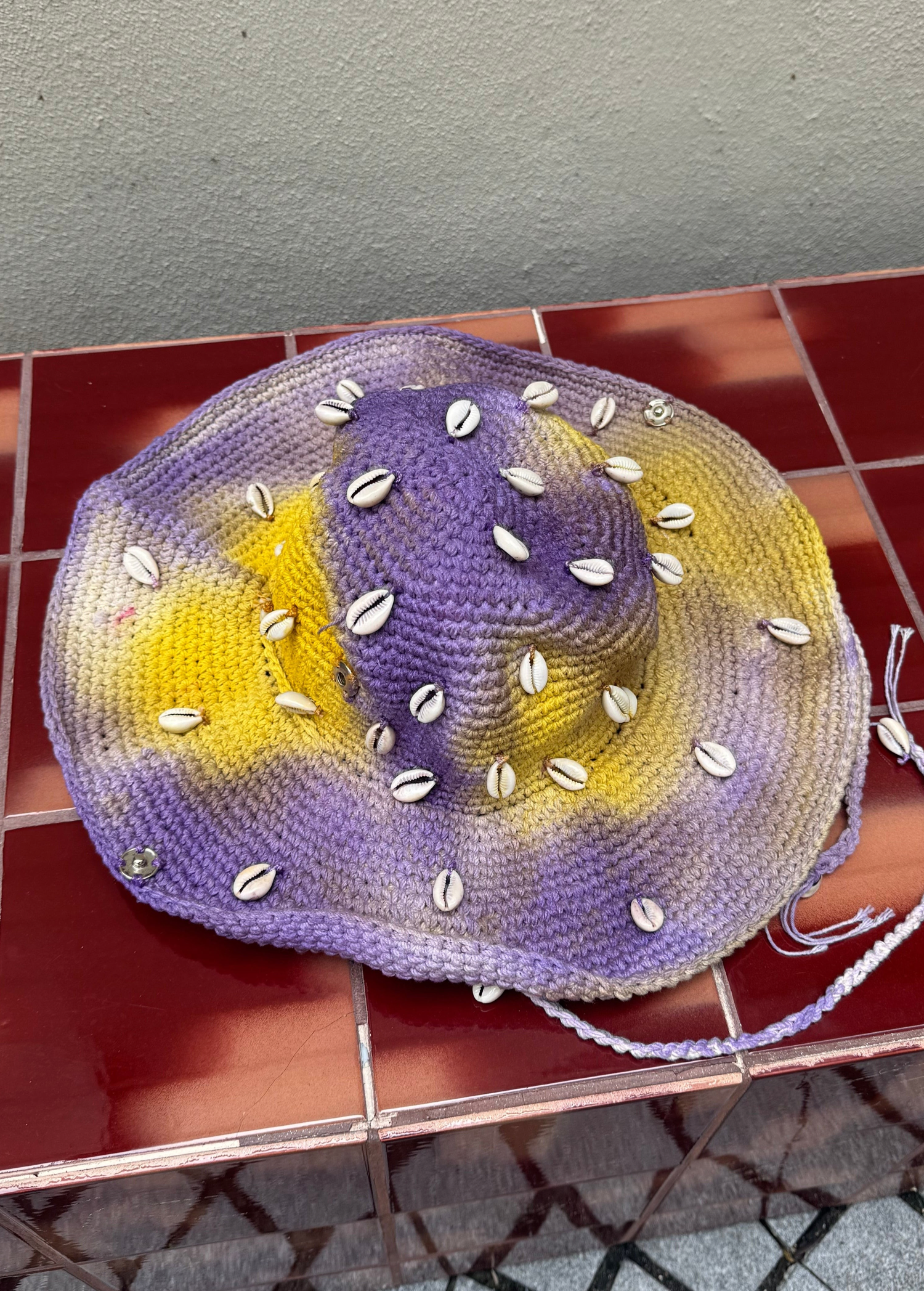 KABE SHOP INC crochet tie-dye hat in purple and yellow with decorative white seashells and a braided chin strap.