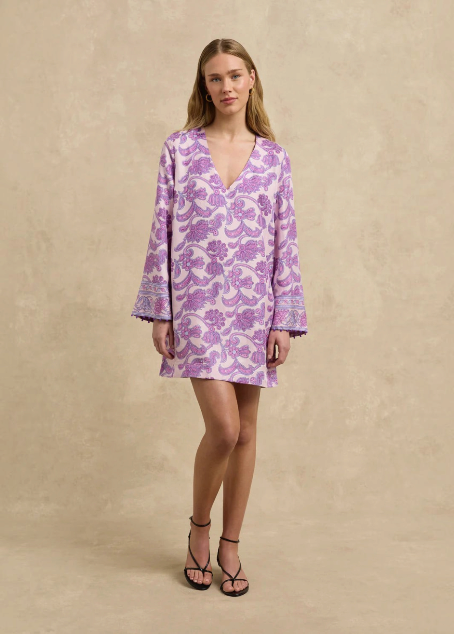 Model wearing a KABE SHOP INC Jaya tunic dress in light pink with a purple paisley print and long flared sleeves.