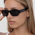 A person with straight, light brown hair wears the Jordyn black festival sunglasses and a white top, holding one arm of the sunglasses. The background is plain and light-colored.