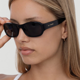 A person with straight, light brown hair wears the Jordyn black festival sunglasses and a white top, holding one arm of the sunglasses. The background is plain and light-colored.