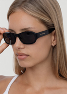 A person with straight, light brown hair wears the Jordyn black festival sunglasses and a white top, holding one arm of the sunglasses. The background is plain and light-colored.
