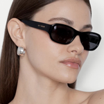 A woman with straight brown hair wears the Jordyn rectangular black festival sunglasses, which offer UV protection, and large silver earrings. She has a neutral expression and is photographed against a plain light background.