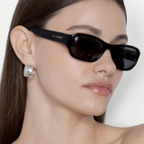 A woman with straight brown hair wears the Jordyn rectangular black festival sunglasses, which offer UV protection, and large silver earrings. She has a neutral expression and is photographed against a plain light background.