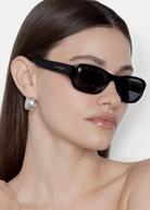 A woman with straight brown hair wears the Jordyn rectangular black festival sunglasses, which offer UV protection, and large silver earrings. She has a neutral expression and is photographed against a plain light background.