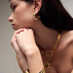 Woman with long dark hair wearing the Kaia Earrings—gold plated hoops—styled with a chunky gold chain necklace featuring a heart pendant, a gold cuff bracelet, and a red top, posed with her hands near her chin looking to the side.