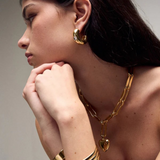 Woman with long dark hair wearing the Kaia Earrings—gold plated hoops—styled with a chunky gold chain necklace featuring a heart pendant, a gold cuff bracelet, and a red top, posed with her hands near her chin looking to the side.