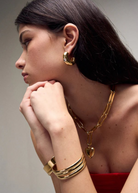 Woman with long dark hair wearing the Kaia Earrings—gold plated hoops—styled with a chunky gold chain necklace featuring a heart pendant, a gold cuff bracelet, and a red top, posed with her hands near her chin looking to the side.
