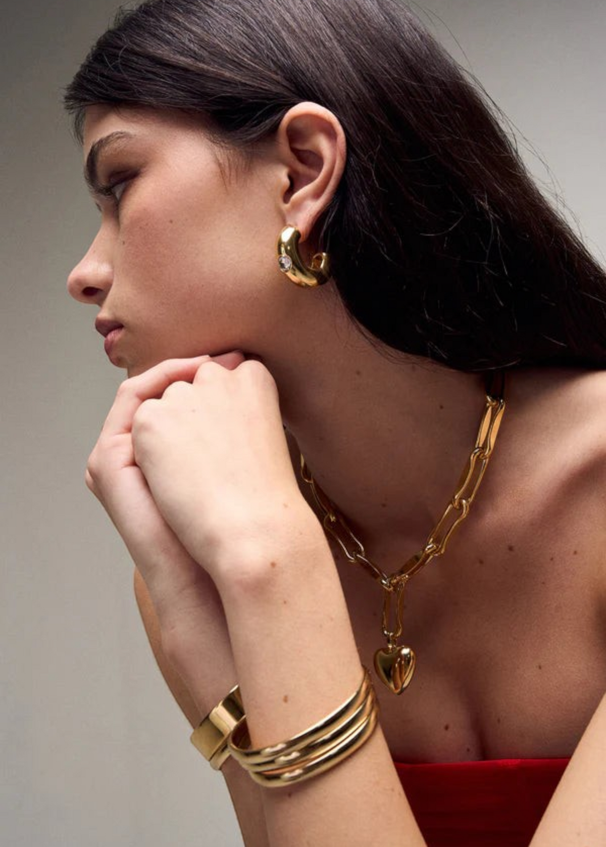 Woman with long dark hair wearing the Kaia Earrings—gold plated hoops—styled with a chunky gold chain necklace featuring a heart pendant, a gold cuff bracelet, and a red top, posed with her hands near her chin looking to the side.