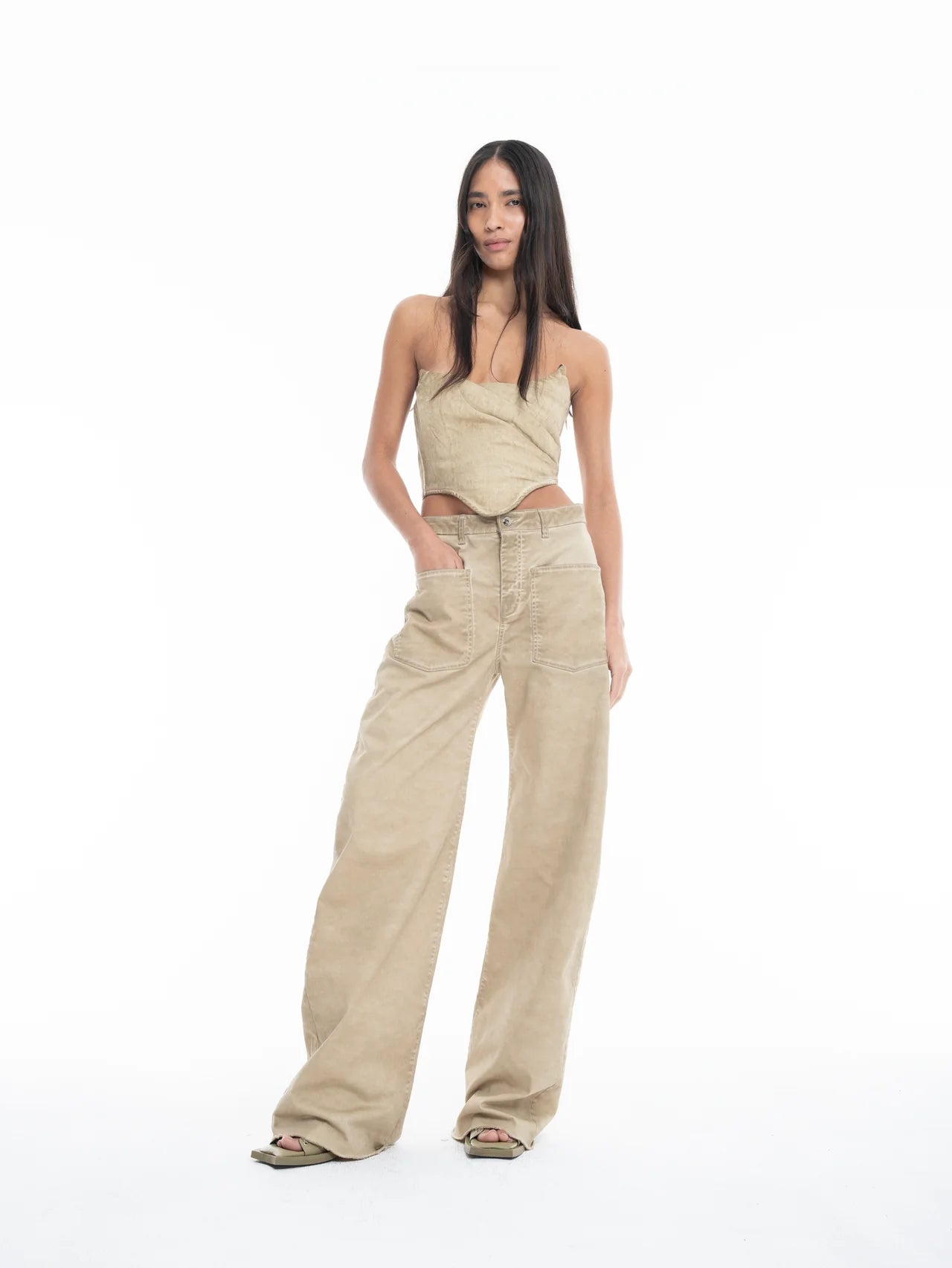 A woman with long dark hair wears a beige, asymmetrical sleeveless top and Cold Dye Boyfriend Jeans with Patch Pockets. She stands confidently against a plain white background, with one hand in her patch pocket and the other by her side.