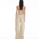 A woman stands facing away against a plain white background, wearing a strapless beige corset top and the Cold Dye Boyfriend Jeans with Patch Pockets, featuring a loose, wide-leg khaki design and a yellow tag visible in the back patch pocket.
