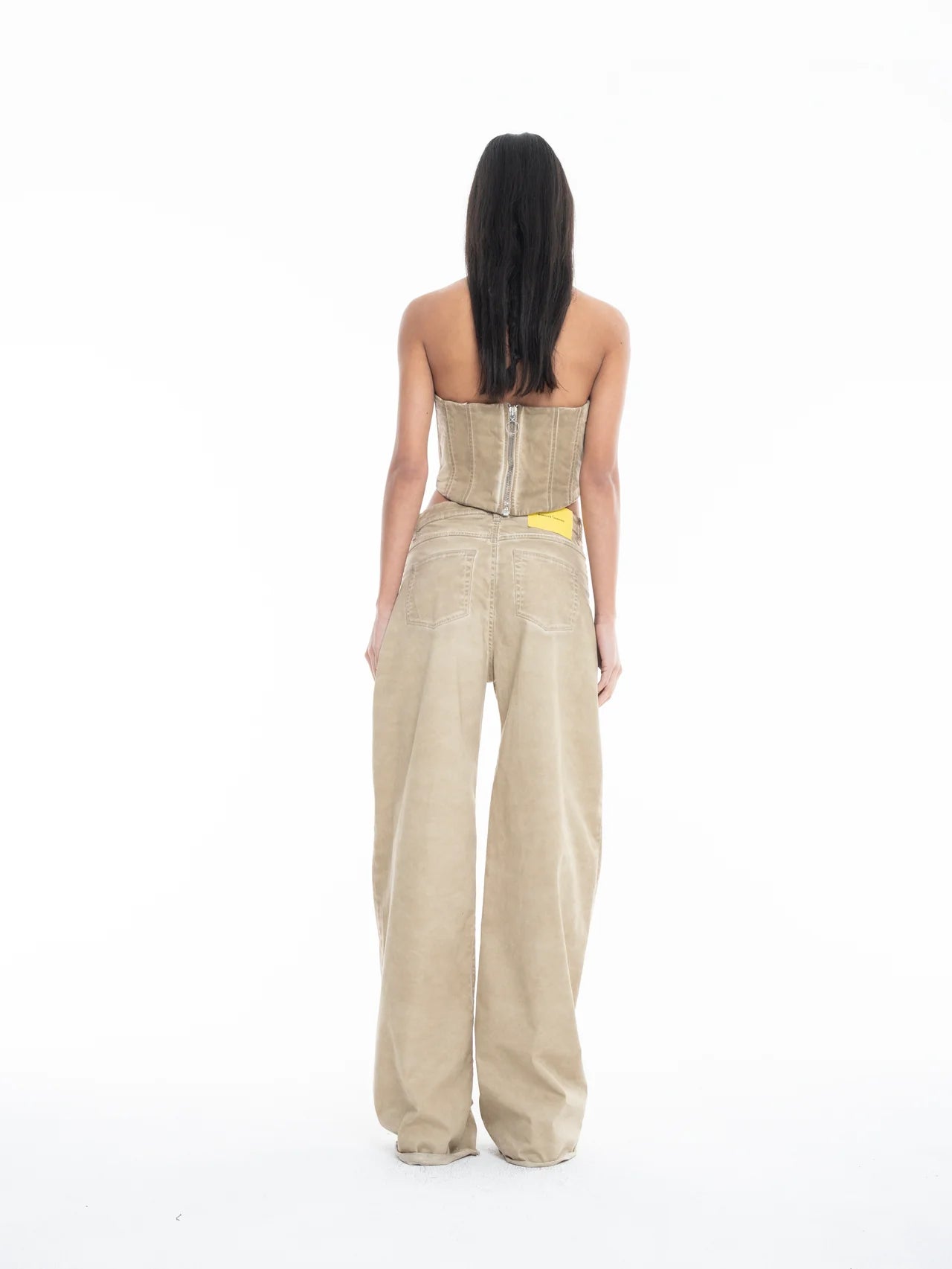 A woman stands facing away against a plain white background, wearing a strapless beige corset top and the Cold Dye Boyfriend Jeans with Patch Pockets, featuring a loose, wide-leg khaki design and a yellow tag visible in the back patch pocket.