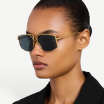 Wearing the gold-plated, oversized-frame Miranda Sunglasses and a black blazer, a person with slicked-back hair poses against a plain light background.