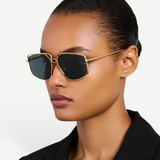 Wearing the gold-plated, oversized-frame Miranda Sunglasses and a black blazer, a person with slicked-back hair poses against a plain light background.