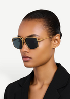 Wearing the gold-plated, oversized-frame Miranda Sunglasses and a black blazer, a person with slicked-back hair poses against a plain light background.