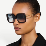Wearing the Macy Sunglasses—featuring oversized square frames crafted from tortoiseshell acetate and dark gradient lenses—a person pairs them with a sleek black blazer while posing against a plain light background.