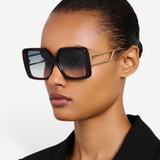 Wearing the Macy Sunglasses—featuring oversized square frames crafted from tortoiseshell acetate and dark gradient lenses—a person pairs them with a sleek black blazer while posing against a plain light background.