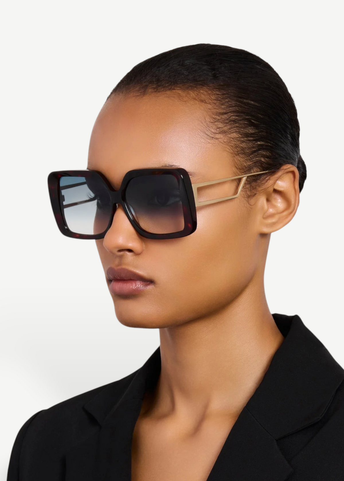 Wearing the Macy Sunglasses—featuring oversized square frames crafted from tortoiseshell acetate and dark gradient lenses—a person pairs them with a sleek black blazer while posing against a plain light background.