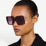A woman with slicked-back hair wears the Macy Sunglasses, oversized square frames crafted from luxurious Italian acetate in maroon, accented by gold-plated temples, styled with a black blazer against a plain light background.