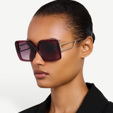 A woman with slicked-back hair wears the Macy Sunglasses, oversized square frames crafted from luxurious Italian acetate in maroon, accented by gold-plated temples, styled with a black blazer against a plain light background.