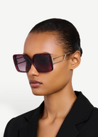A woman with slicked-back hair wears the Macy Sunglasses, oversized square frames crafted from luxurious Italian acetate in maroon, accented by gold-plated temples, styled with a black blazer against a plain light background.