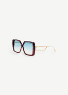 The Macy Sunglasses feature oversized square frames crafted from red tortoiseshell acetate, paired with thin gold arms and aqua gradient lenses, displayed on a white background.