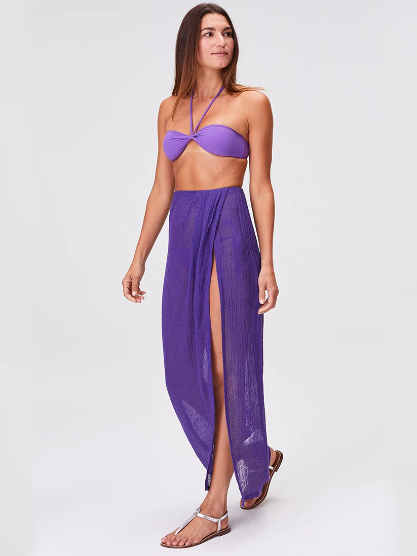 A woman wearing a purple bandeau bikini top and the Sarong Skirt in Purple Blueberry Sorrento Gauze with a high slit, accessorized with silver sandals, stands on a plain light background.