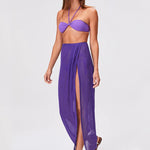 A woman wearing a purple bandeau bikini top and the Sarong Skirt in Purple Blueberry Sorrento Gauze with a high slit, accessorized with silver sandals, stands on a plain light background.