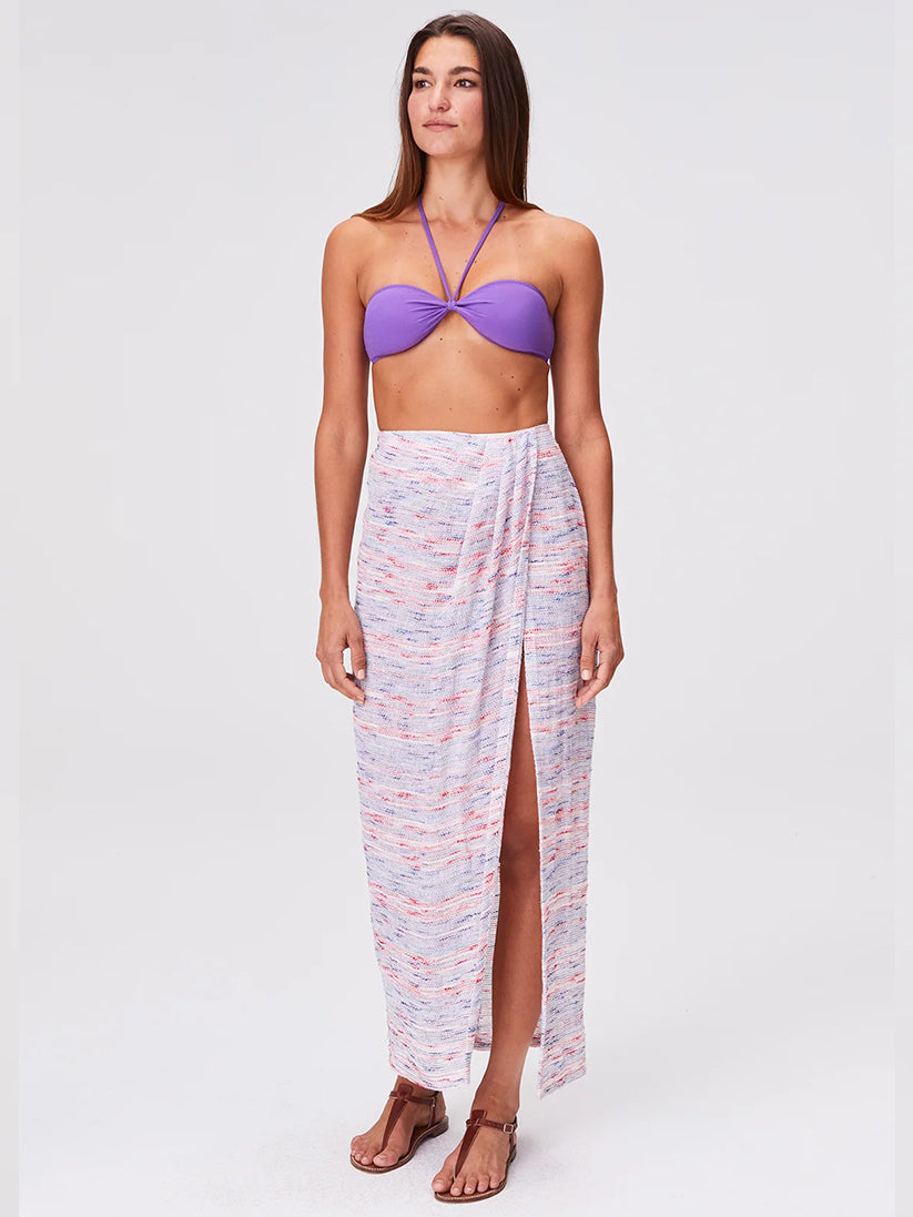 A woman stands wearing a purple bandeau bikini top, the Sarong Skirt—an airy long white gauze skirt with multi-colored stripes and a front slit—and brown sandals against a plain light background for an effortlessly chic beachwear look.