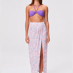 A woman stands wearing a purple bandeau bikini top, the Sarong Skirt—an airy long white gauze skirt with multi-colored stripes and a front slit—and brown sandals against a plain light background for an effortlessly chic beachwear look.