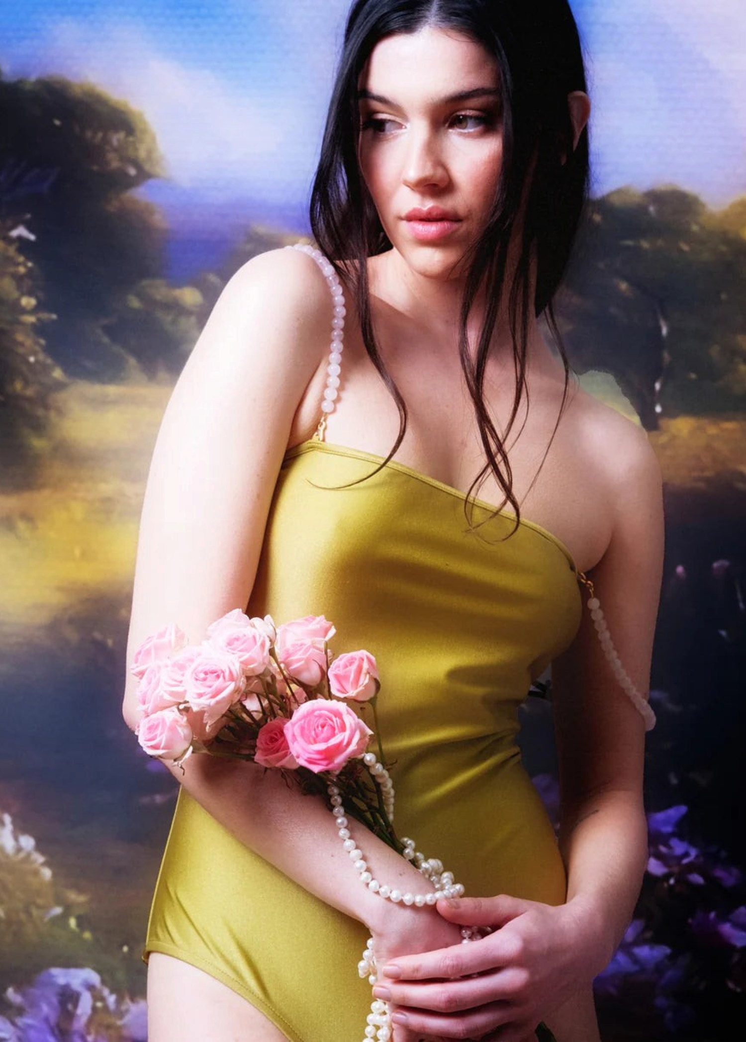 Wearing a yellow dress, a woman holds pink roses and pearl strands, standing before a painted backdrop of greenery and sky, her look elevated by the handmade-in-Paris Petra Straps.