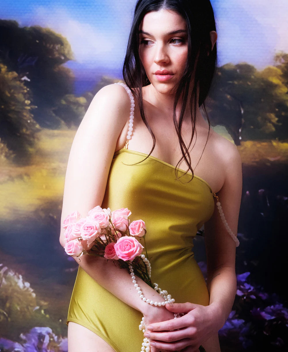 Wearing a yellow dress, a woman holds pink roses and pearl strands, standing before a painted backdrop of greenery and sky, her look elevated by the handmade-in-Paris Petra Straps.
