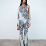 Woman standing against a plain white wall, wearing the Maha Top—a sheer, one-shoulder top with a black and white zebra print—styled with an oversized fit and accessorized with bracelets and rings.