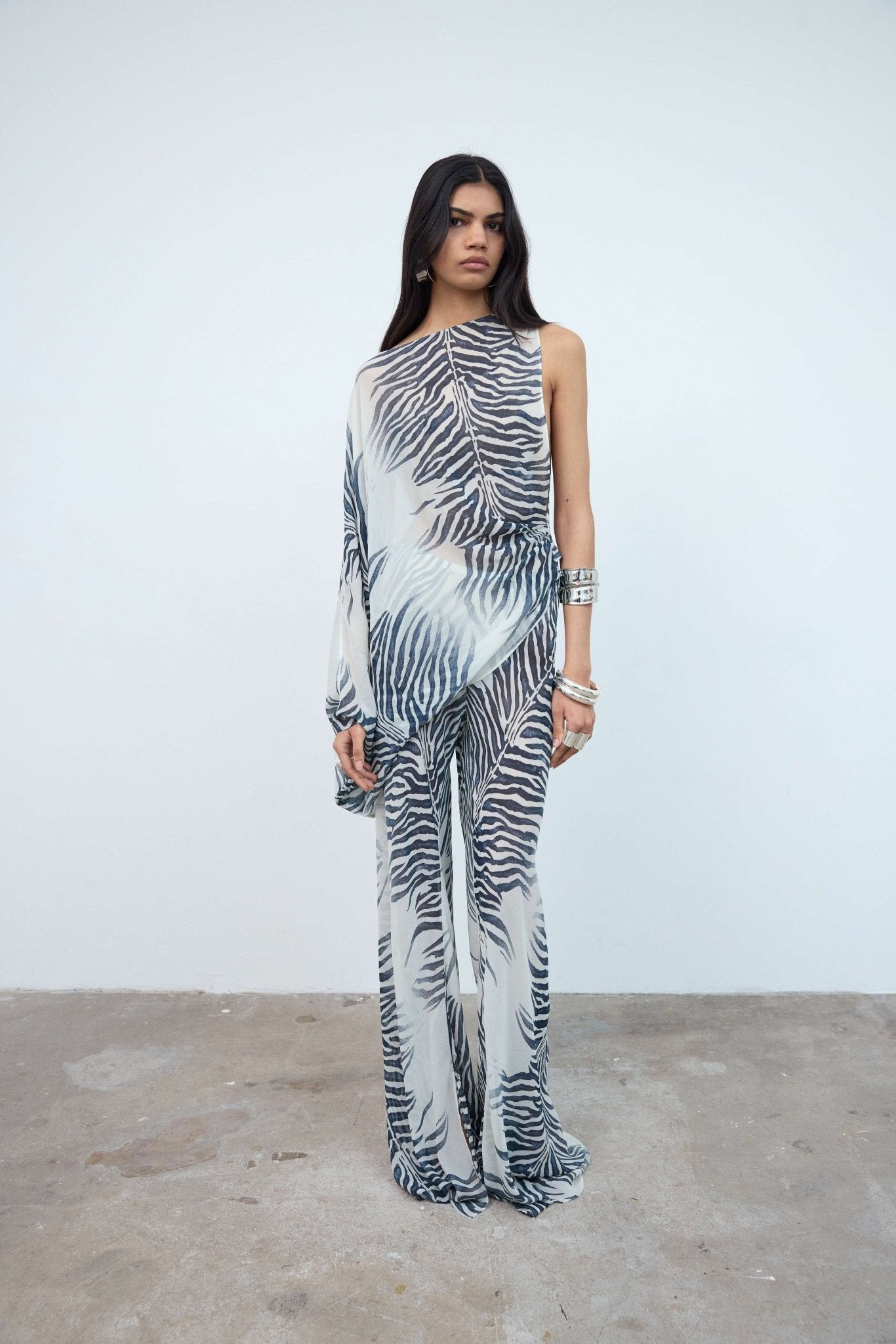 Woman standing against a plain white wall, wearing the Maha Top—a sheer, one-shoulder top with a black and white zebra print—styled with an oversized fit and accessorized with bracelets and rings.