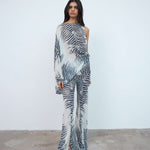 A person stands against a plain white wall, wearing a sheer, one-shouldered jumpsuit with a black and white zebra print pattern and open-toed heels, layered over the oversized fit Maha Top in purple viscose georgette for a bold, contemporary look.