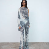 A person stands against a plain white wall, wearing a sheer, one-shouldered jumpsuit with a black and white zebra print pattern and open-toed heels, layered over the oversized fit Maha Top in purple viscose georgette for a bold, contemporary look.
