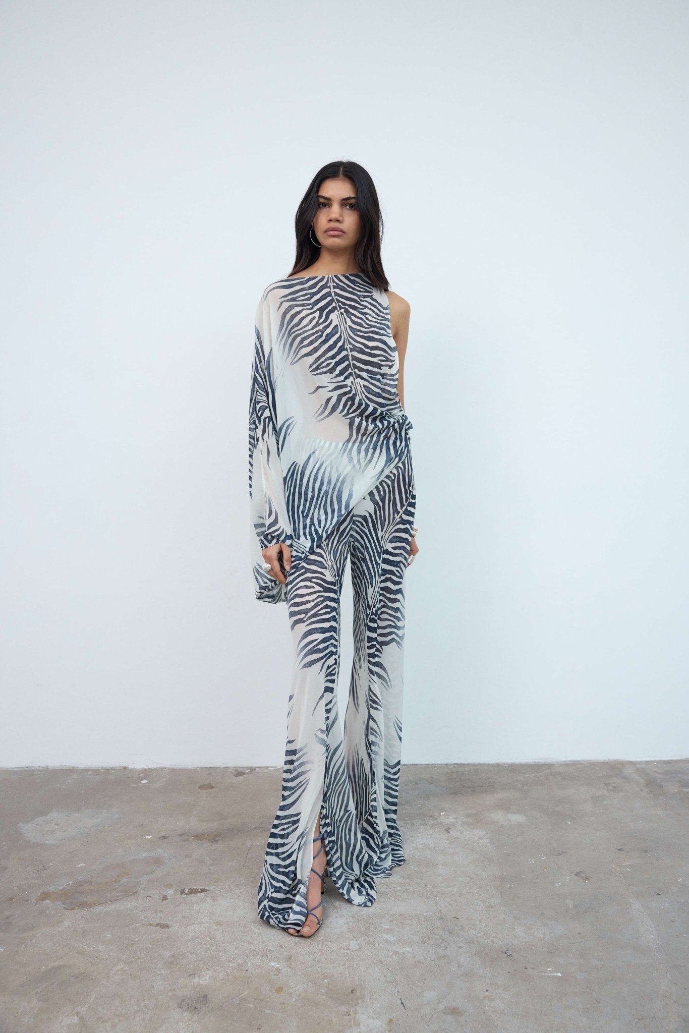 A person stands against a plain white wall, wearing a sheer, one-shouldered jumpsuit with a black and white zebra print pattern and open-toed heels, layered over the oversized fit Maha Top in purple viscose georgette for a bold, contemporary look.