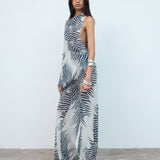 A woman stands against a plain wall wearing a sleeveless, zebra-print maxi dress with side cutouts. She has long dark hair and accessorizes with silver bangles. The concrete floor creates a striking contrast to her bold zebra pattern—a look that echoes the signature statement style of the Luma Trouser.
