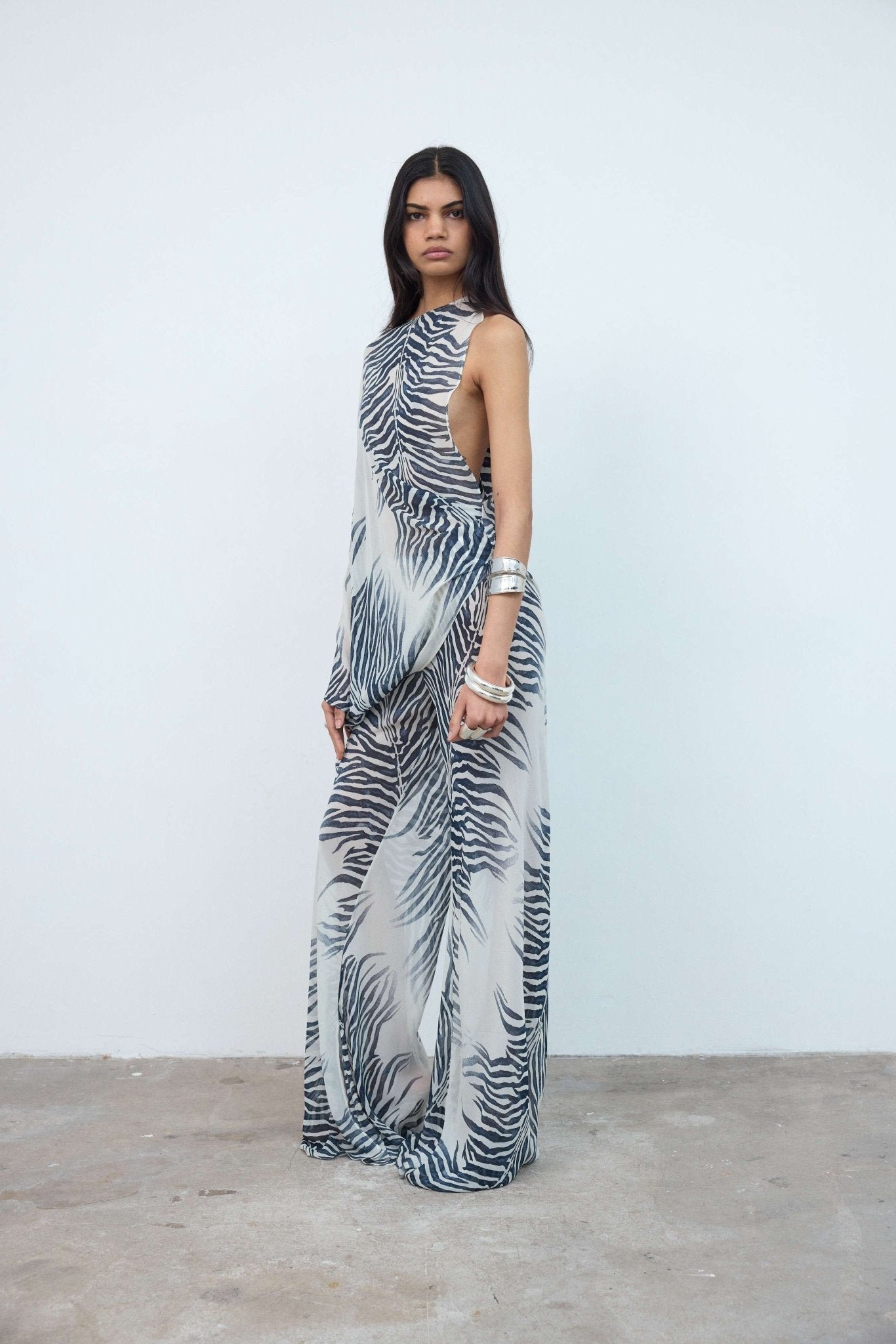 A woman stands against a plain wall wearing a sleeveless, zebra-print maxi dress with side cutouts. She has long dark hair and accessorizes with silver bangles. The concrete floor creates a striking contrast to her bold zebra pattern—a look that echoes the signature statement style of the Luma Trouser.