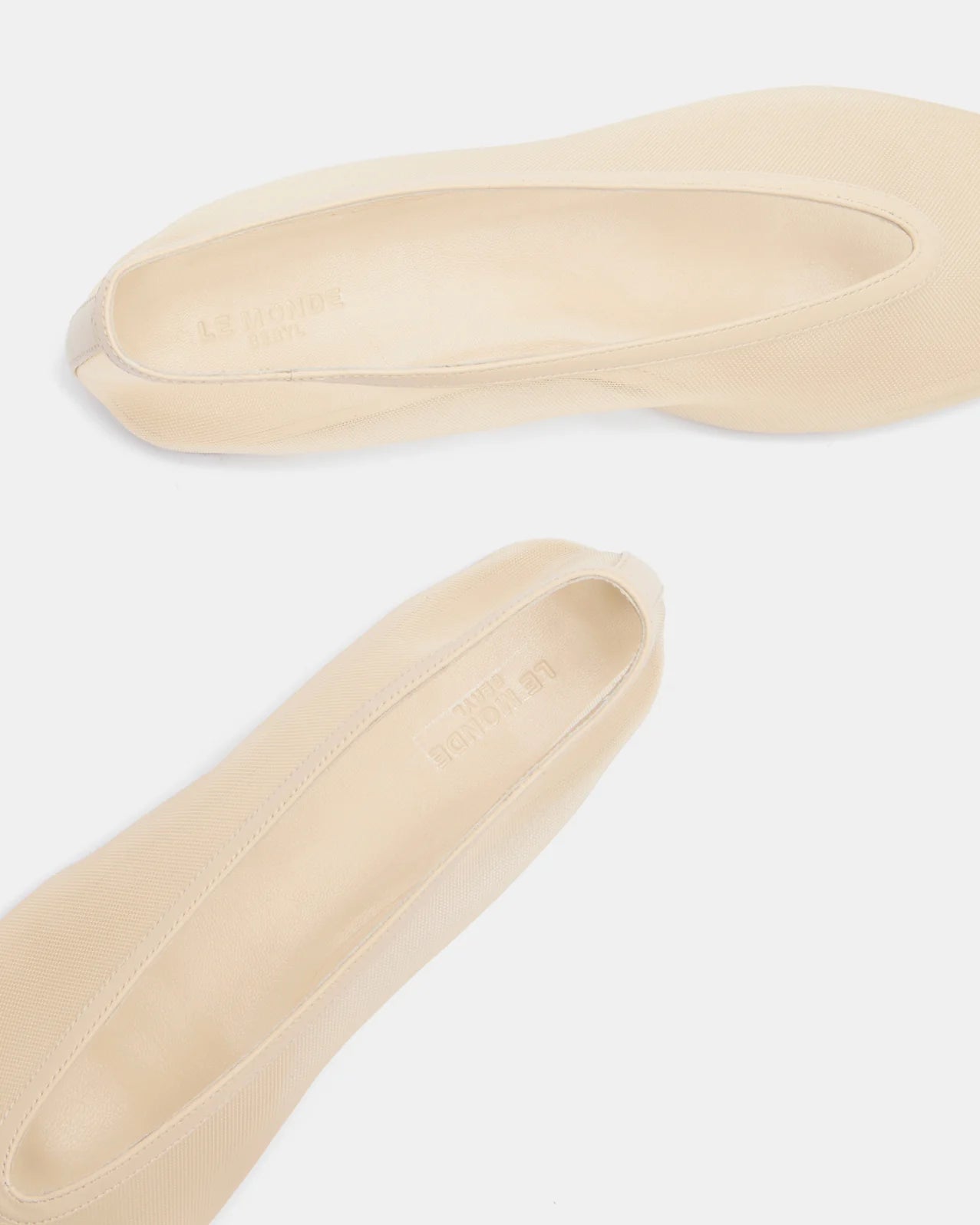 Two beige Luna Slipper Shoes are placed on a white surface, with one shoe positioned above the other. Showcasing a minimalist design and rounded toes, these slip-on Luna Slipper Shoes feature subtle sand mesh detailing for added elegance.