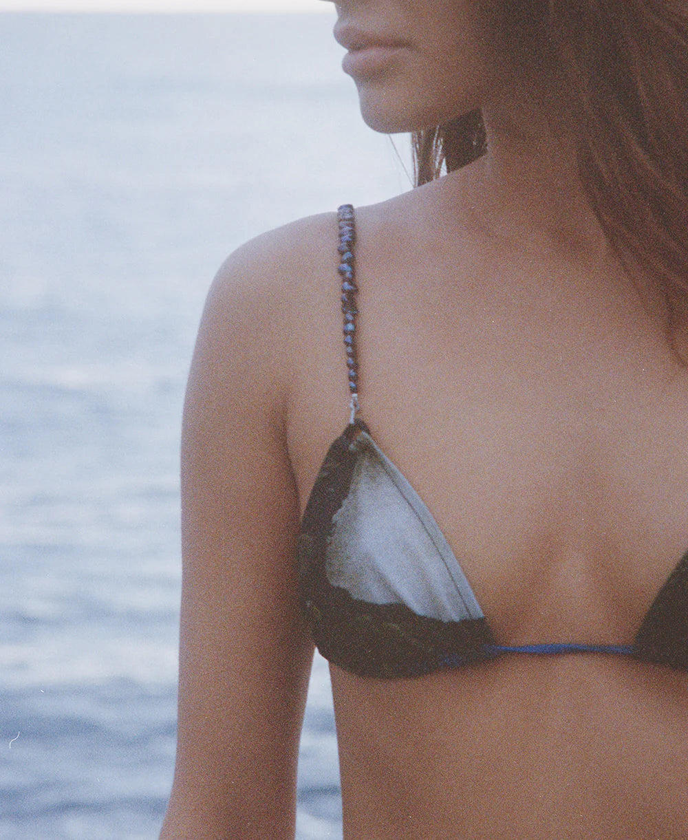 A woman wearing the Dolores Top Classic Swimwear, featuring delicate jewelry straps, stands near the water; her face is partially visible as a calm, blurred ocean forms the background.